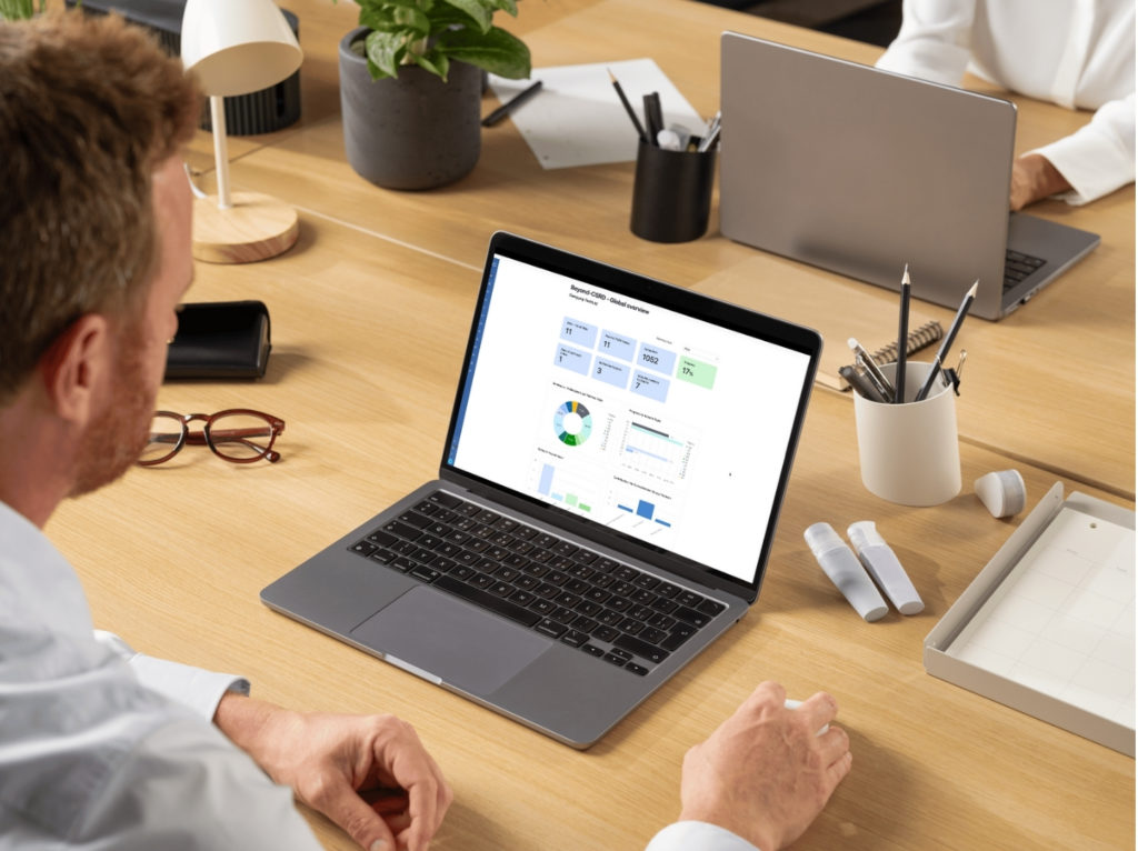 A person seated at a wooden desk looking at a laptop displaying a dashboard of charts and metrics, with office supplies (glasses, pens, markers, potted plant) and a second laptop partially visible in the background.